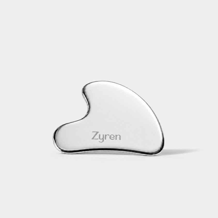 Zyren Gua Sha, a polished metal facial tool engraved with the Zyren logo, lying on white marble